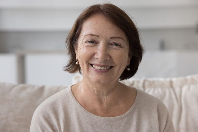 An elderly woman is smiling while sitting on a couch.