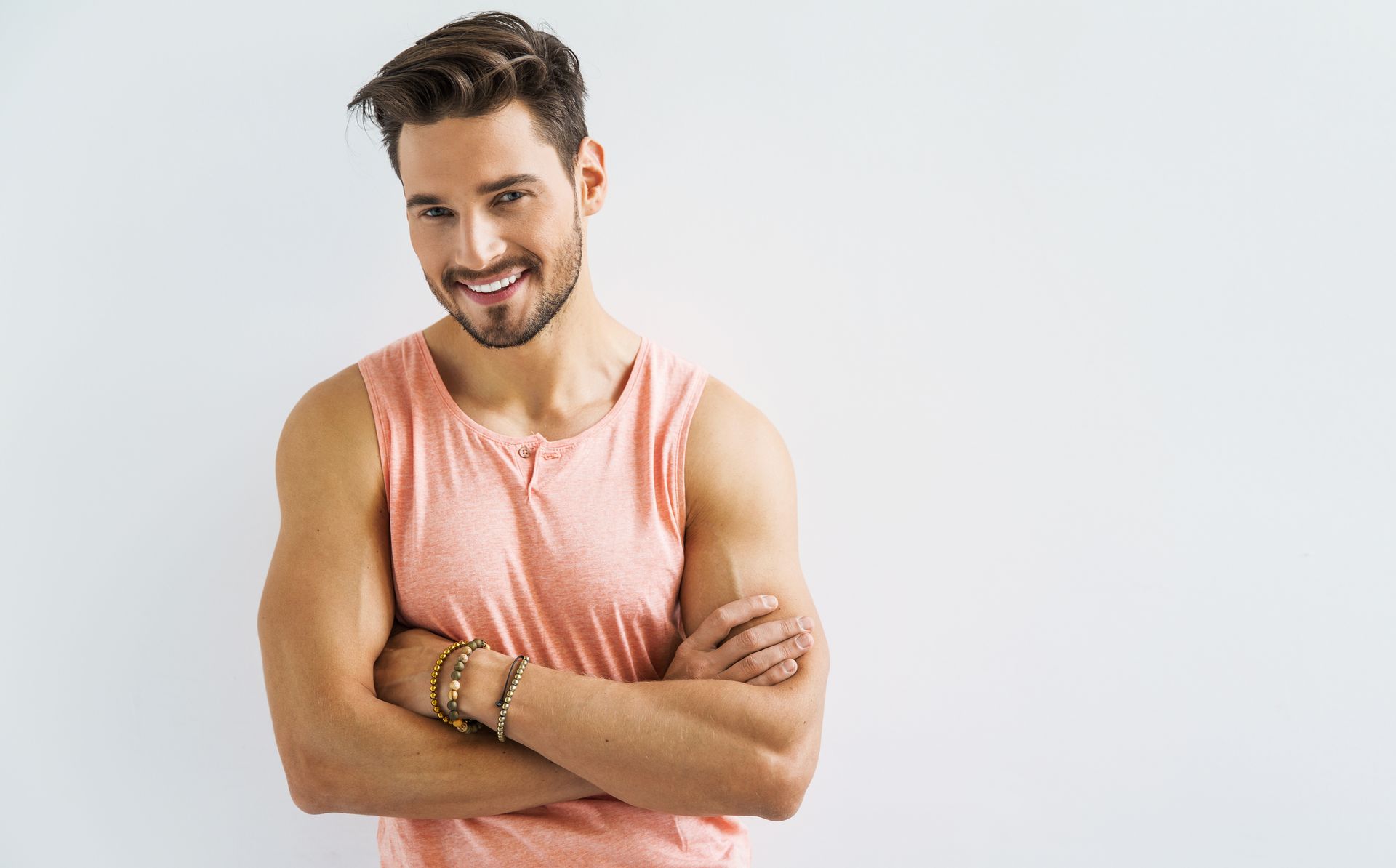 Man with crossed arms smiles, wearing a pink tank top, against a white background.