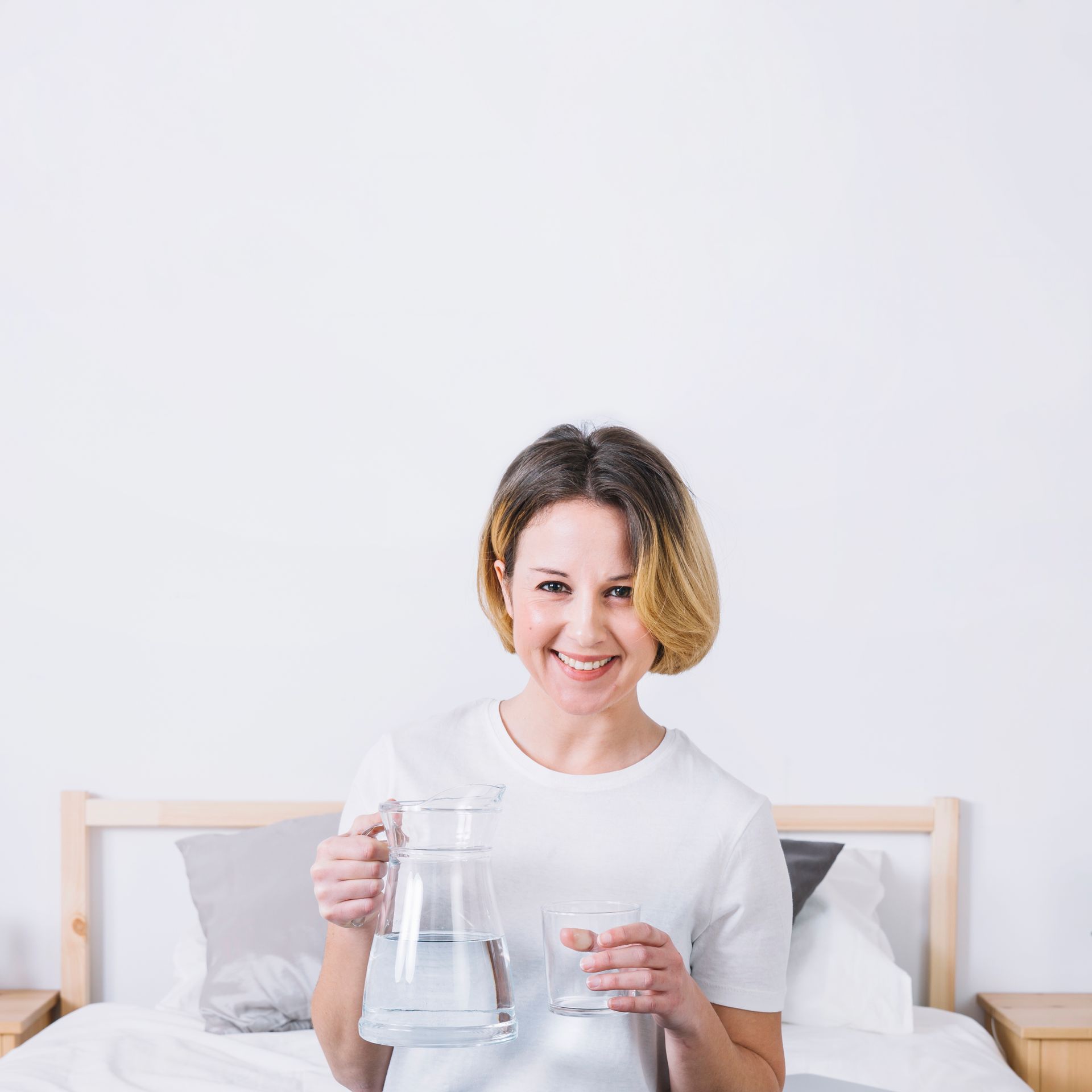 A woman is sitting on a bed holding a pitcher of water and a glass of water.