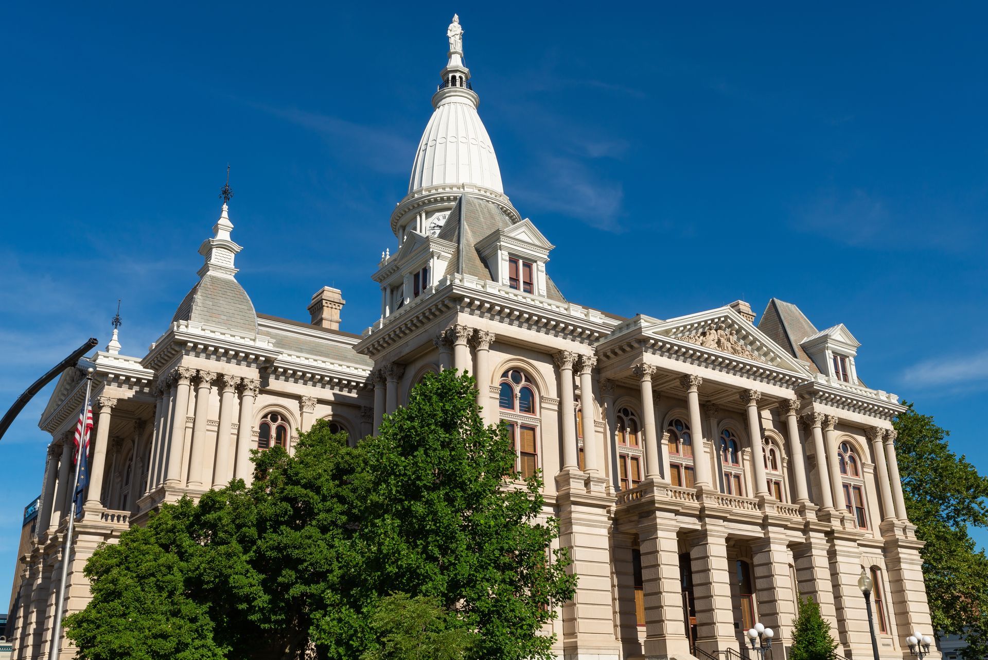 Ornate stone courthouse with a tall white dome against a blue sky, partly framed by green trees