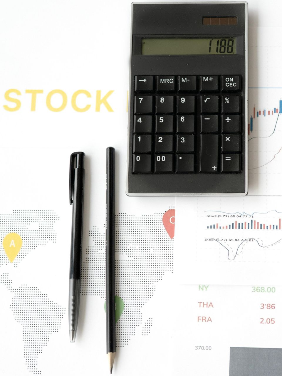 Calculator, pens, and a magnifying glass resting on financial stock market charts and data tables on a white desk.