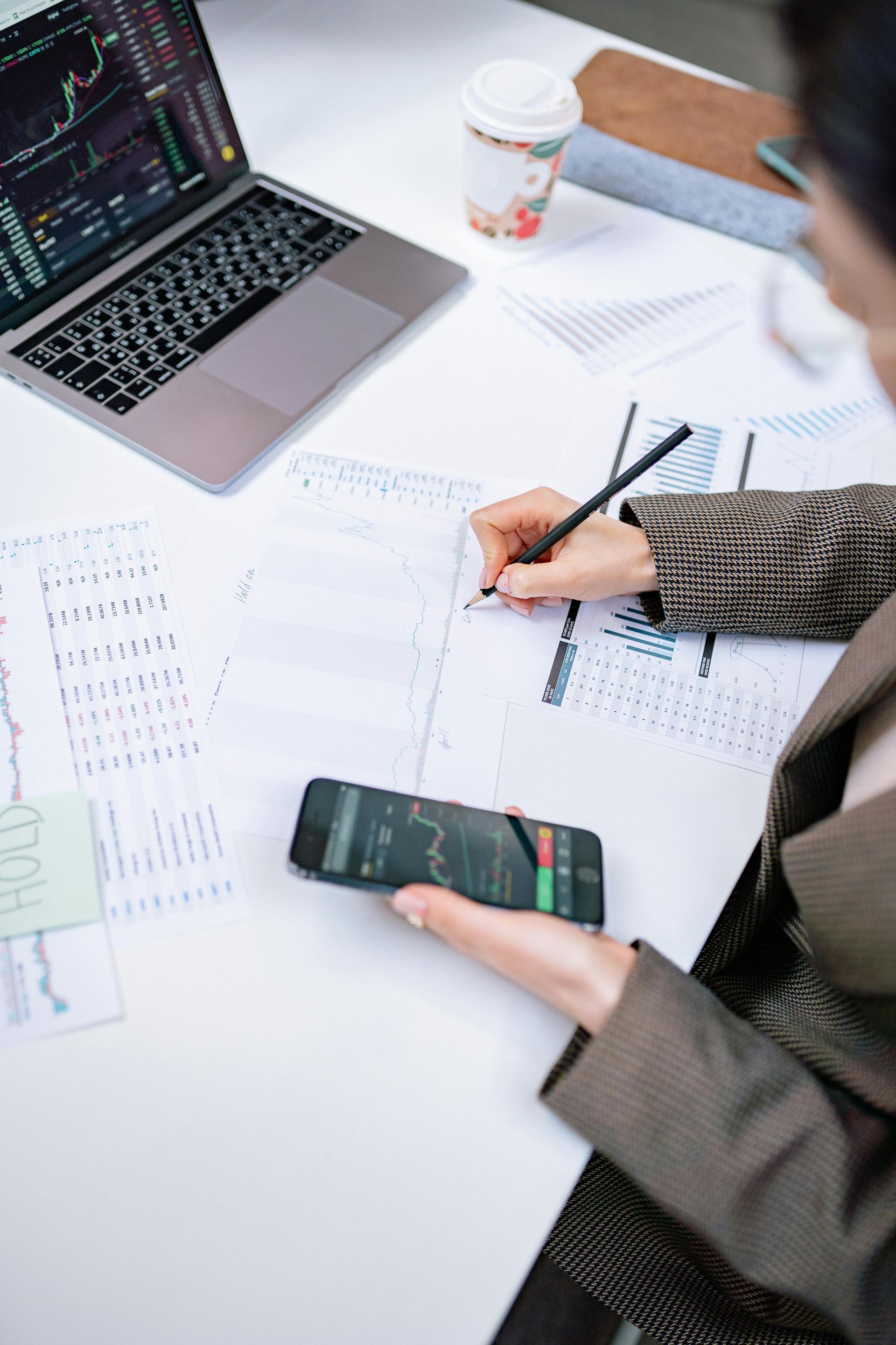 Person writing notes at a desk with a smartphone, laptop, and papers with charts.