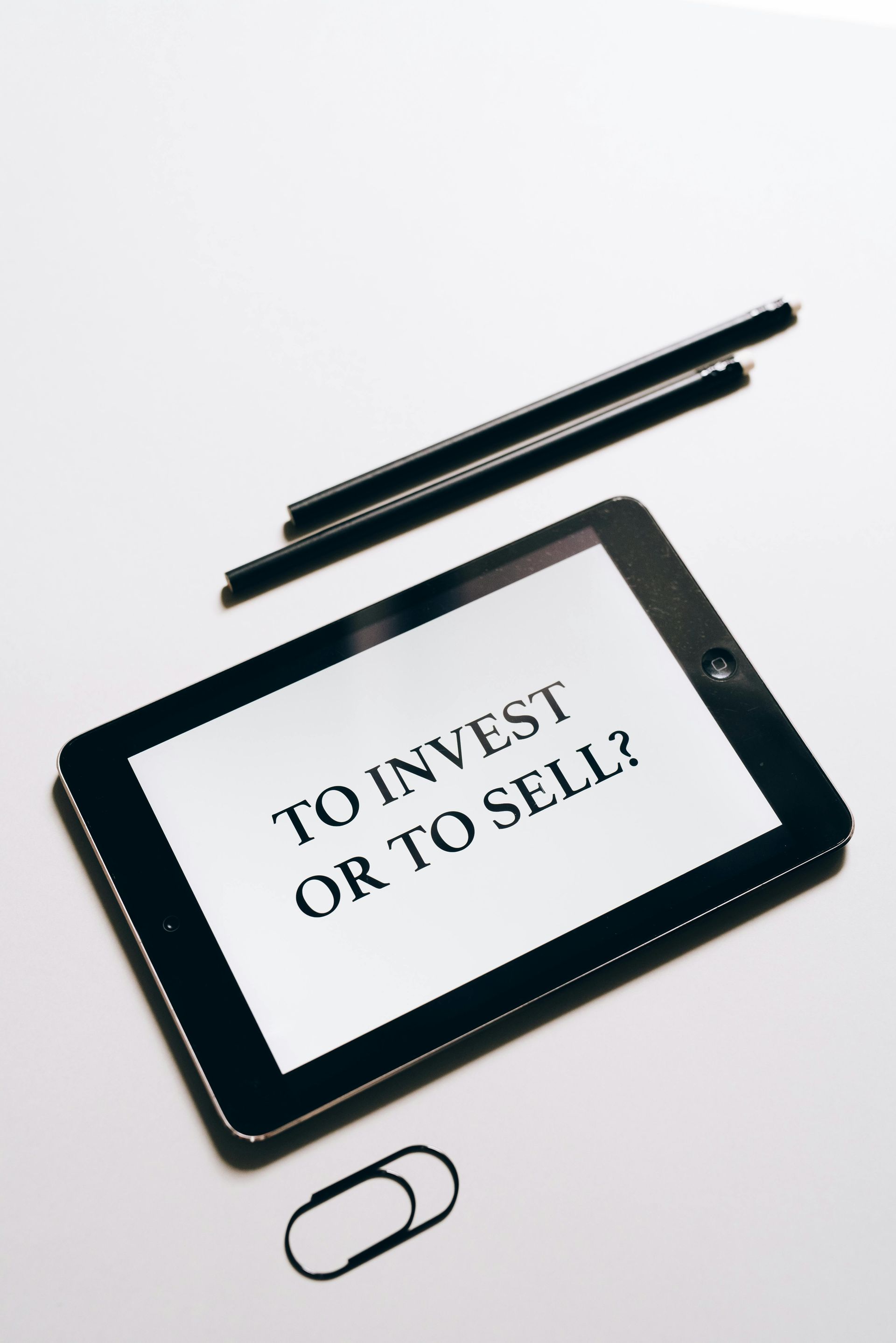 Tablet with “TO INVEST OR TO SELL?” on screen, with stylus and paperclip on a white background