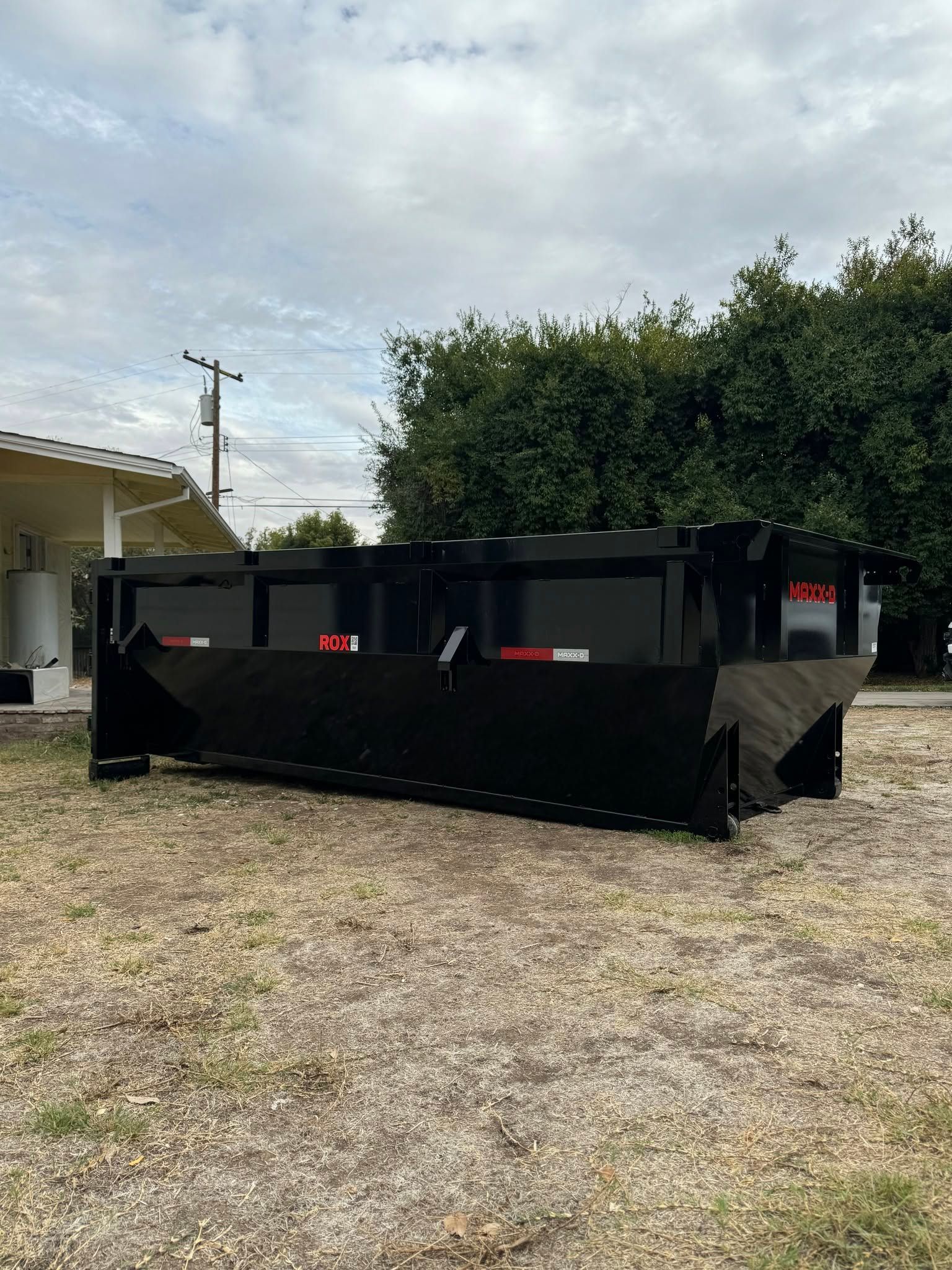 Black construction dumpster on gravel in a yard beside a house and trees