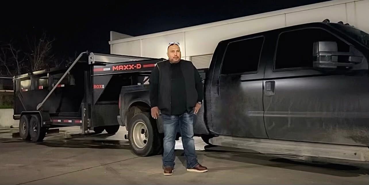 Man standing beside a black truck and trailer at night on a dirt lot
