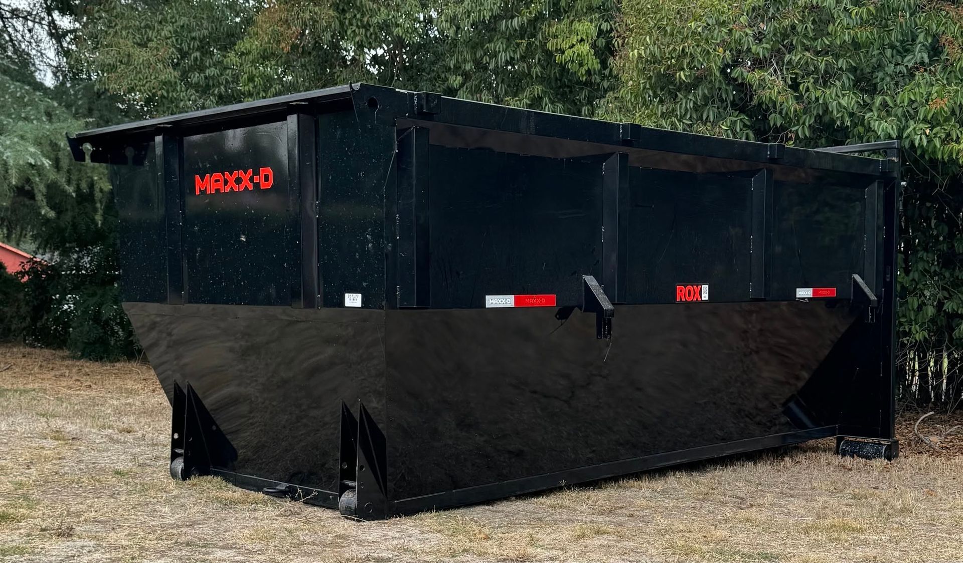 Black industrial dumpster on gravel with red reflective strips, outdoors in front of trees