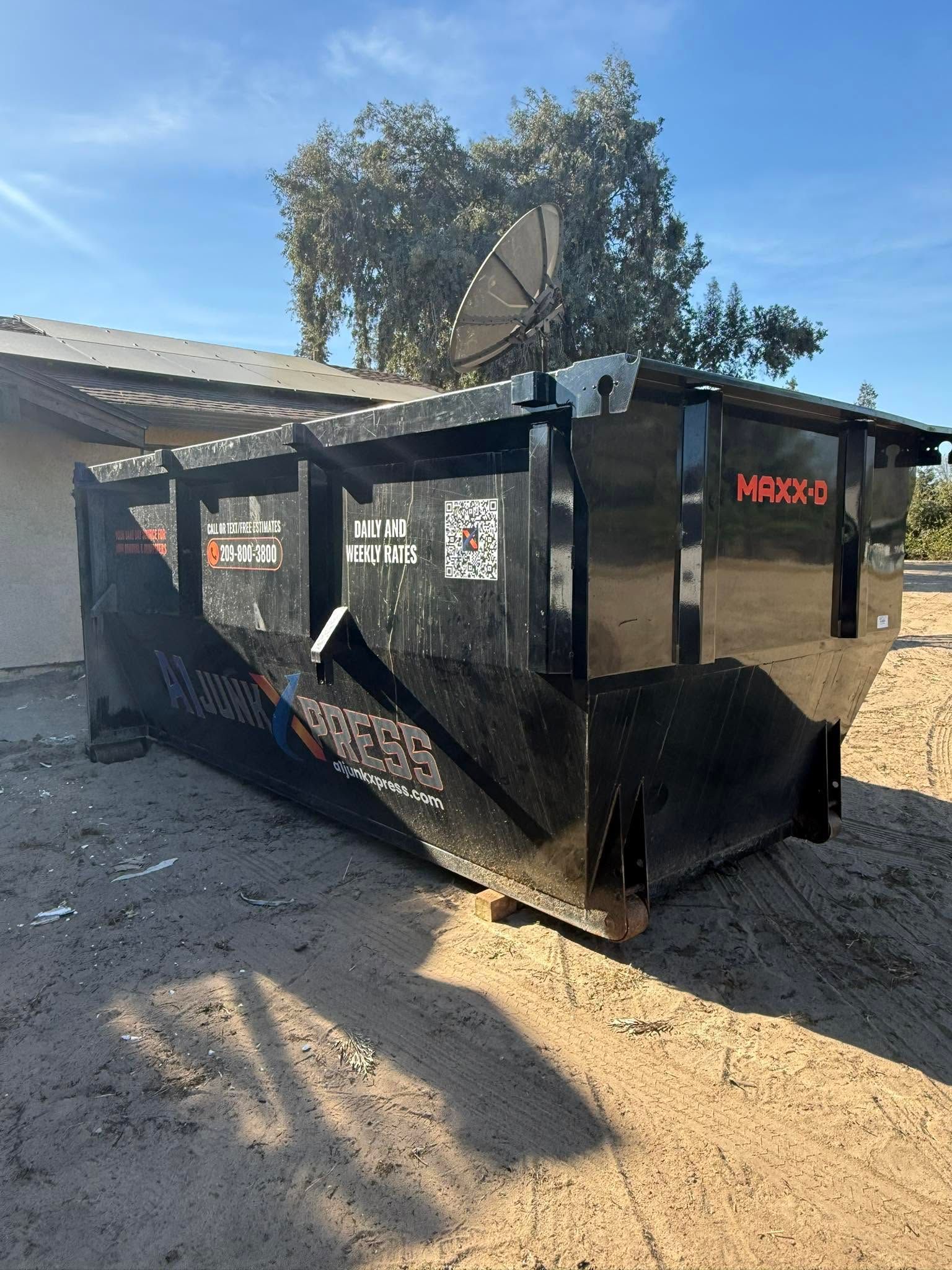 Black industrial dumpster in a gravel lot beside a building, with a satellite dish and tree behind it.