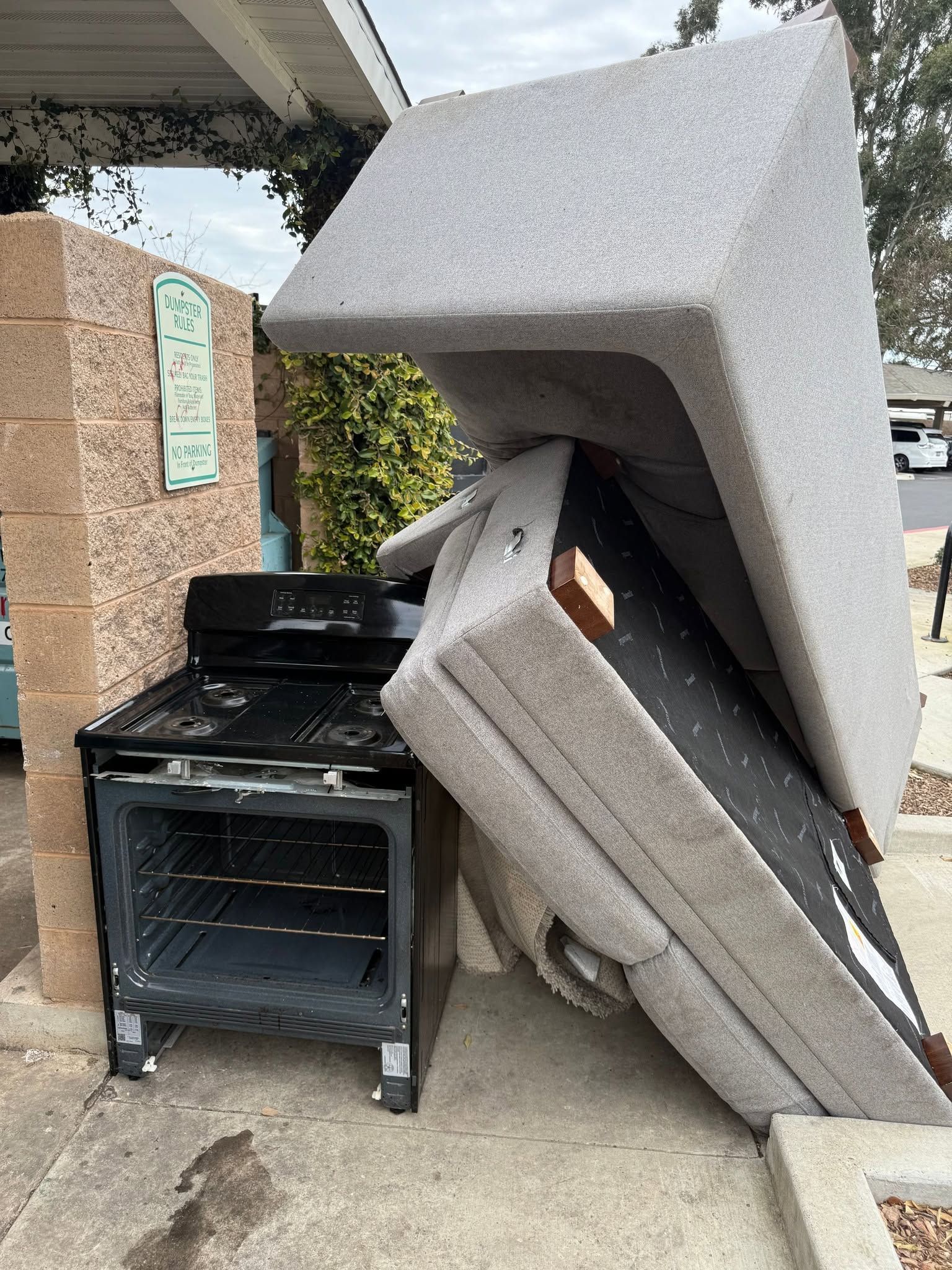 Stove and two foam mattress pieces piled under a carport beside a brick wall