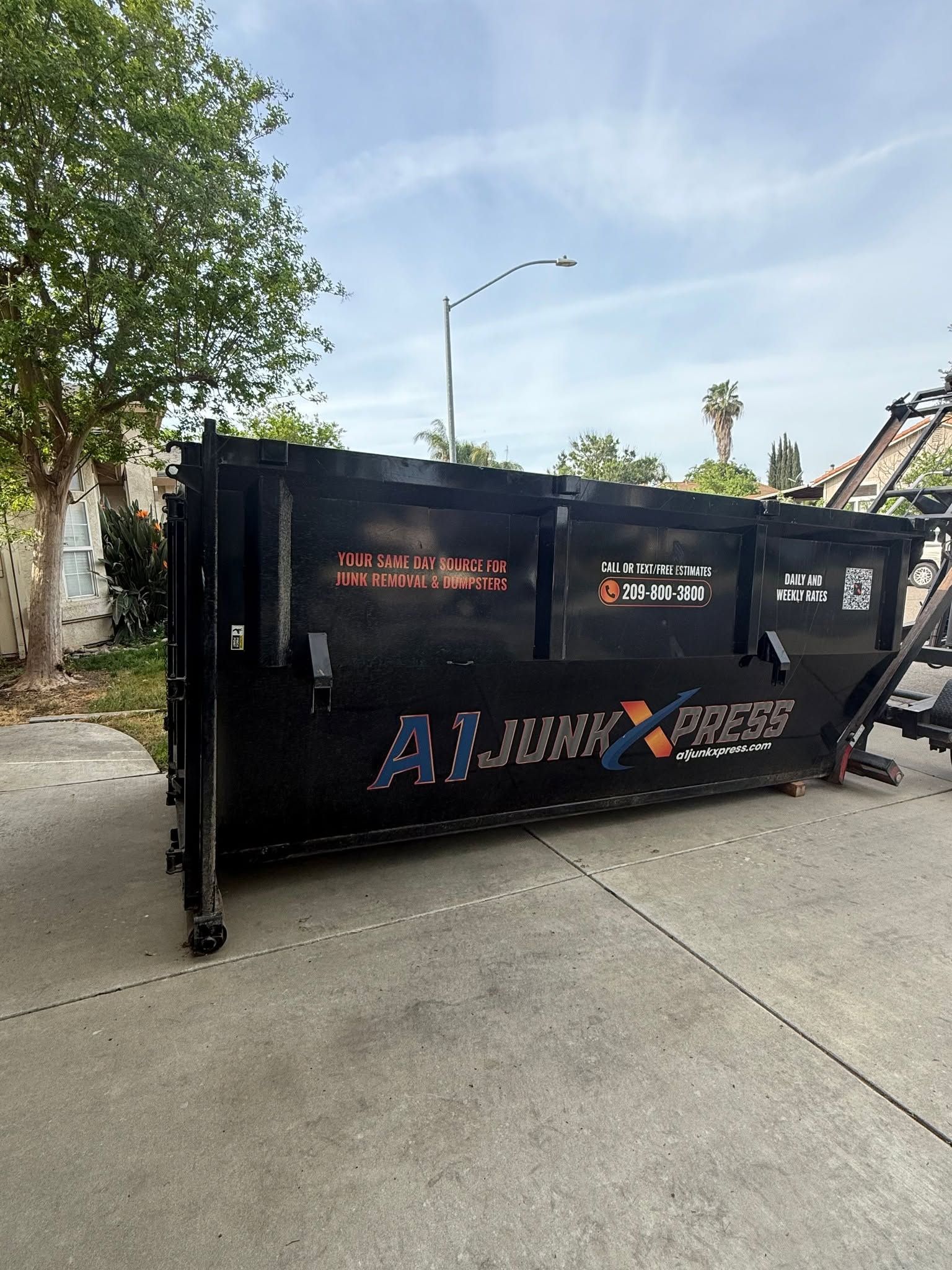 Large black construction dumpster on a concrete street beside a tree and wall.