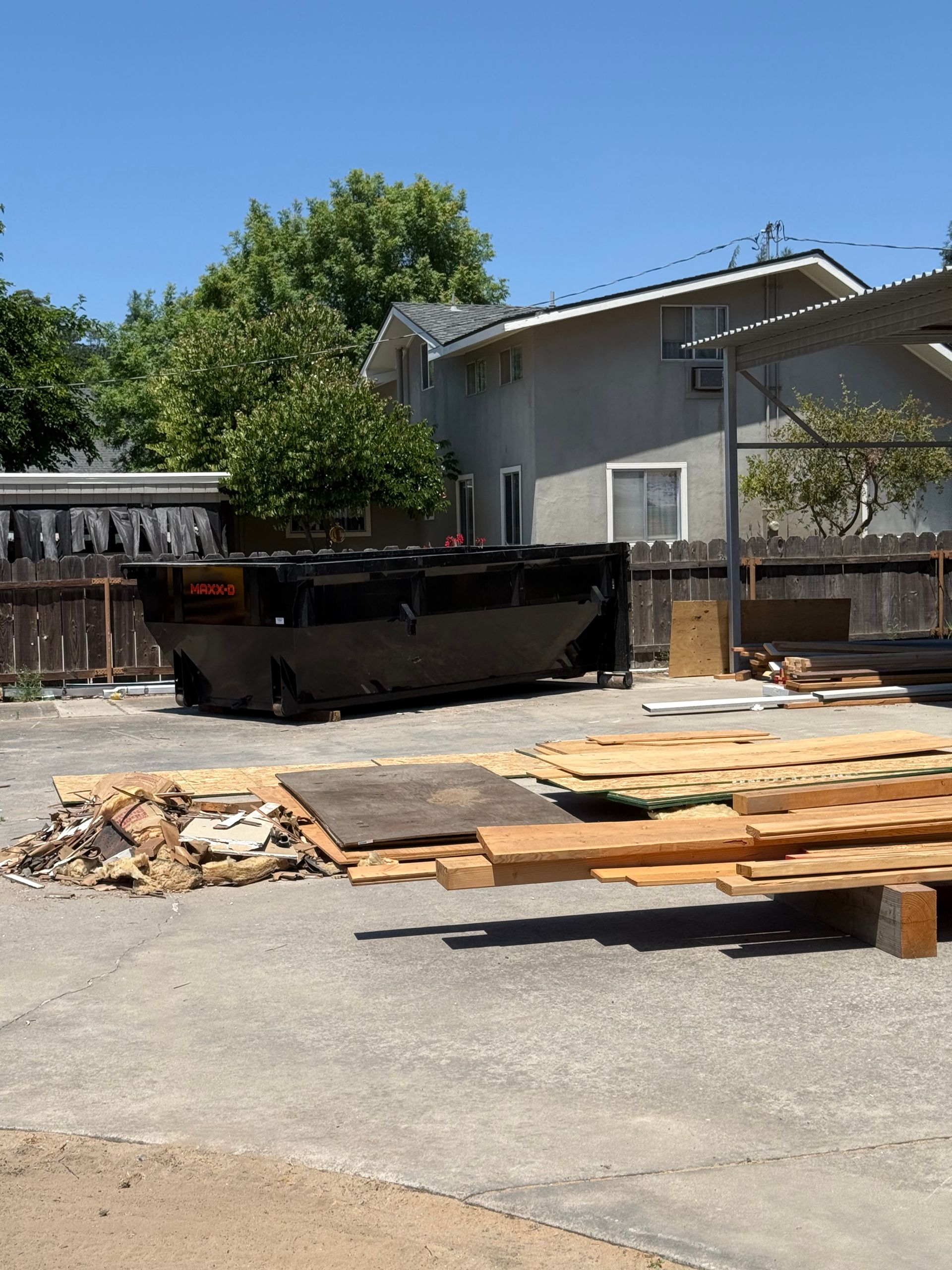 Gray dumpster filled with debris in a suburban driveway beside a house and stacked lumber