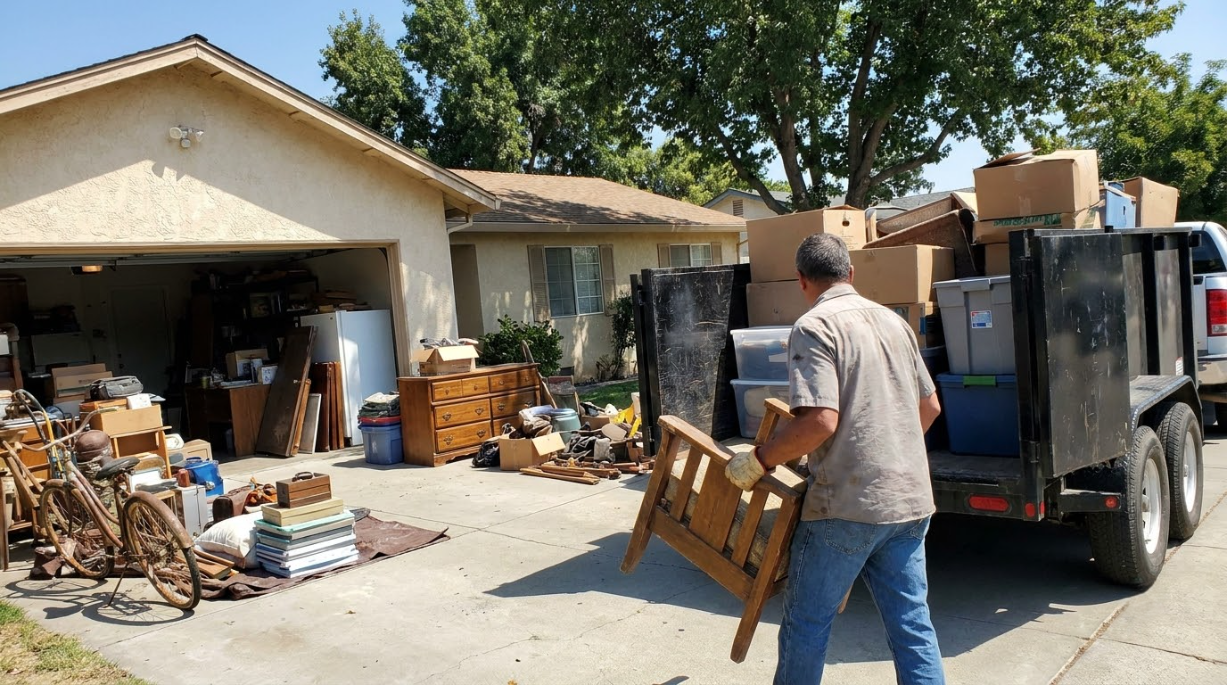 Man unloading furniture and boxes from a van into a cluttered driveway beside an open garage