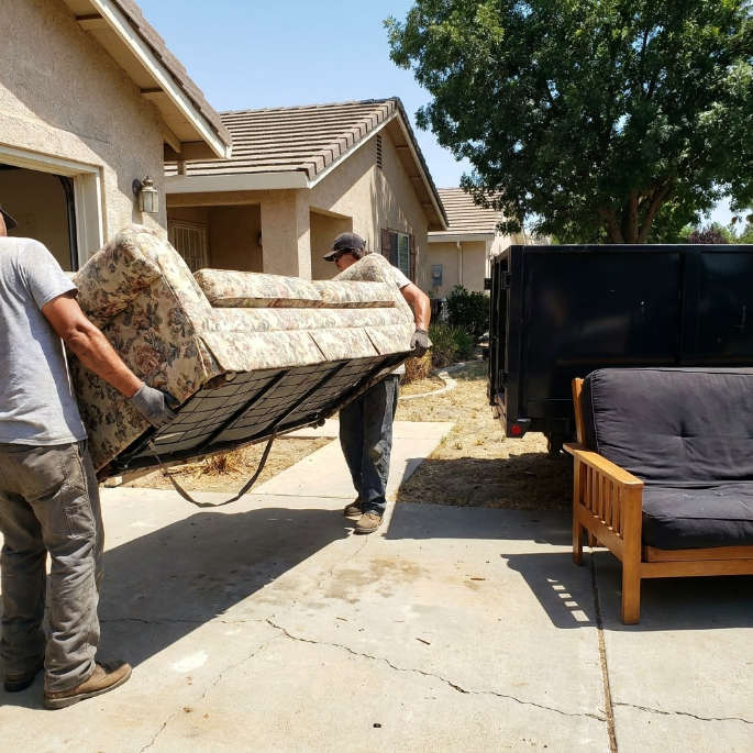 Two people carrying a couch mattress out of a house into a sunny driveway, with a black dump trailer nearby