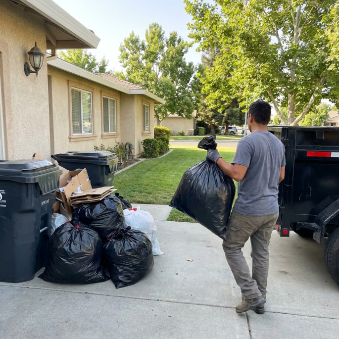 Person carrying trash bags beside a driveway next to a black truck and a house.
