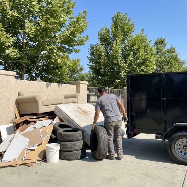 Man loading debris, tires, and an old couch into a black trailer in a sunny driveway.
