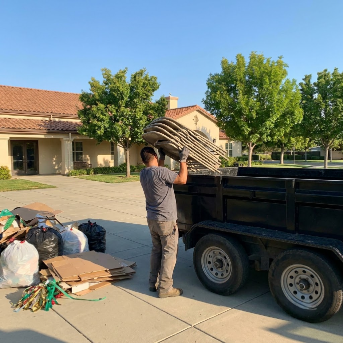 Person loading stacked chairs into a trailer beside piles of household items outside a building