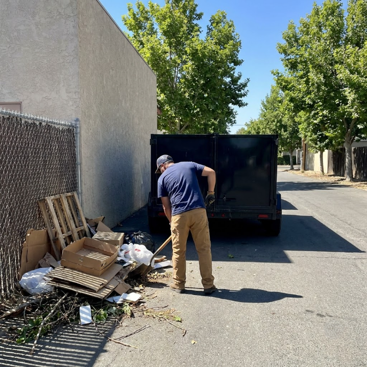 Person loading debris into a black truck beside a wall on a sunny street.