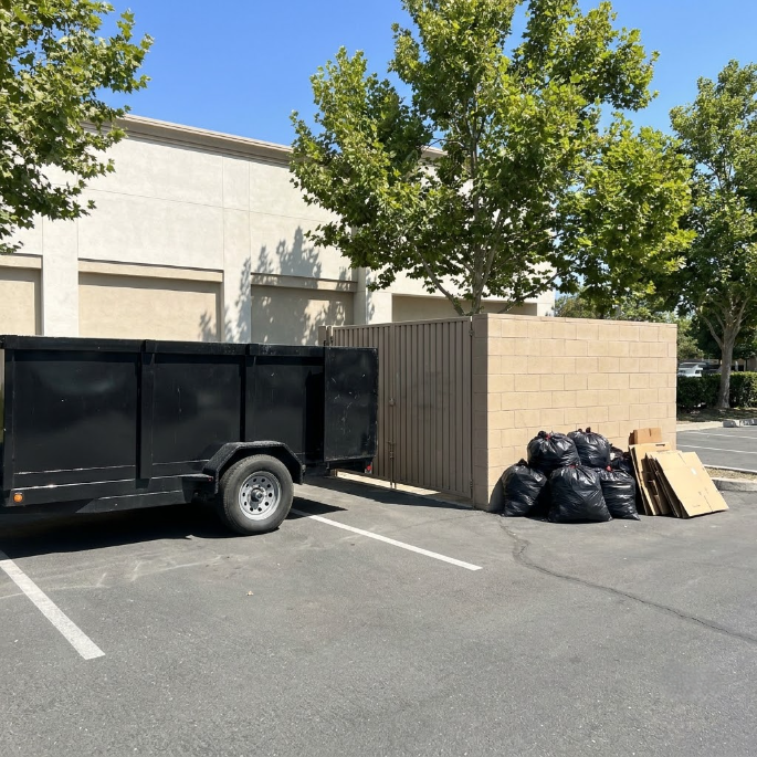 Black utility trailer beside a beige building, with stacked trash bags and cardboard in a sunny parking lot.