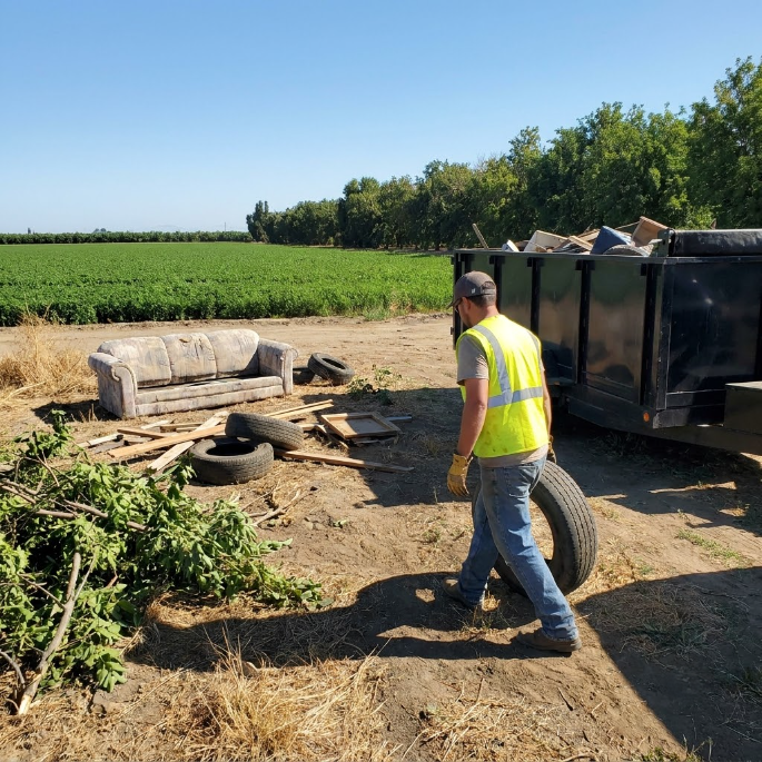 Worker in a neon vest stands beside a dumpster in a dirt field with a couch and debris nearby.