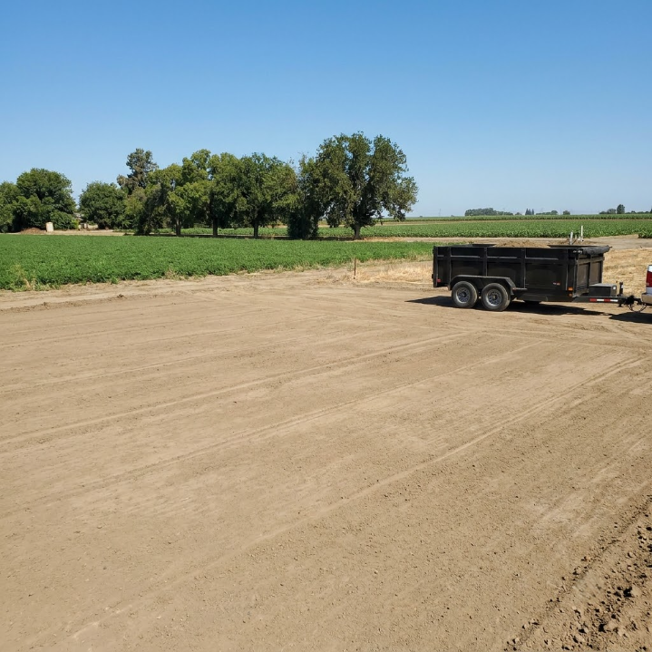 Gravel lot beside green fields and trees under a clear blue sky, with a black trailer on the right.