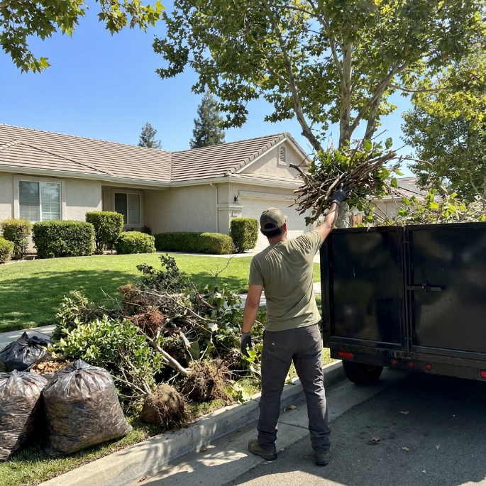 Person loading yard waste into a trailer beside a suburban house on a sunny day