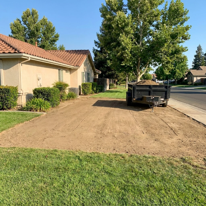 Freshly leveled dirt yard beside a suburban house, with a trailer on a quiet street and trees in back.