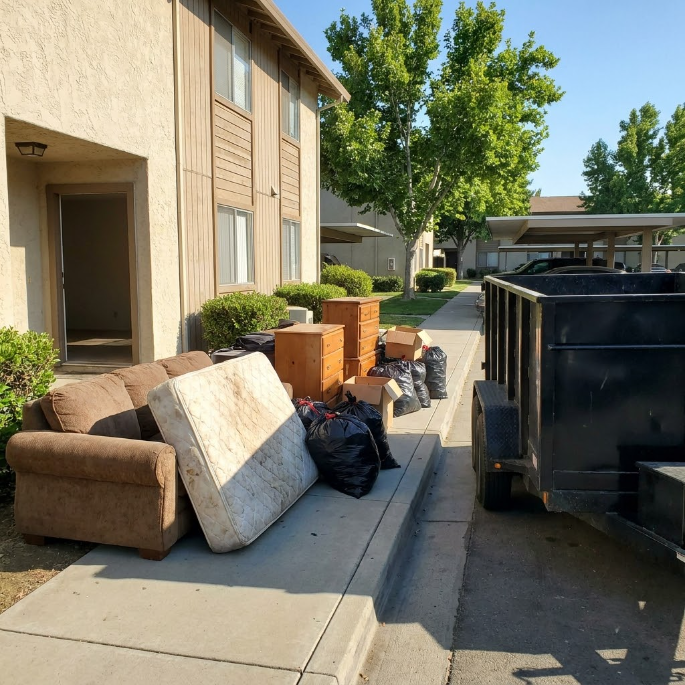 Furniture and trash piled on a curb beside apartment buildings and a dumpster, with mattresses and bags out for pickup