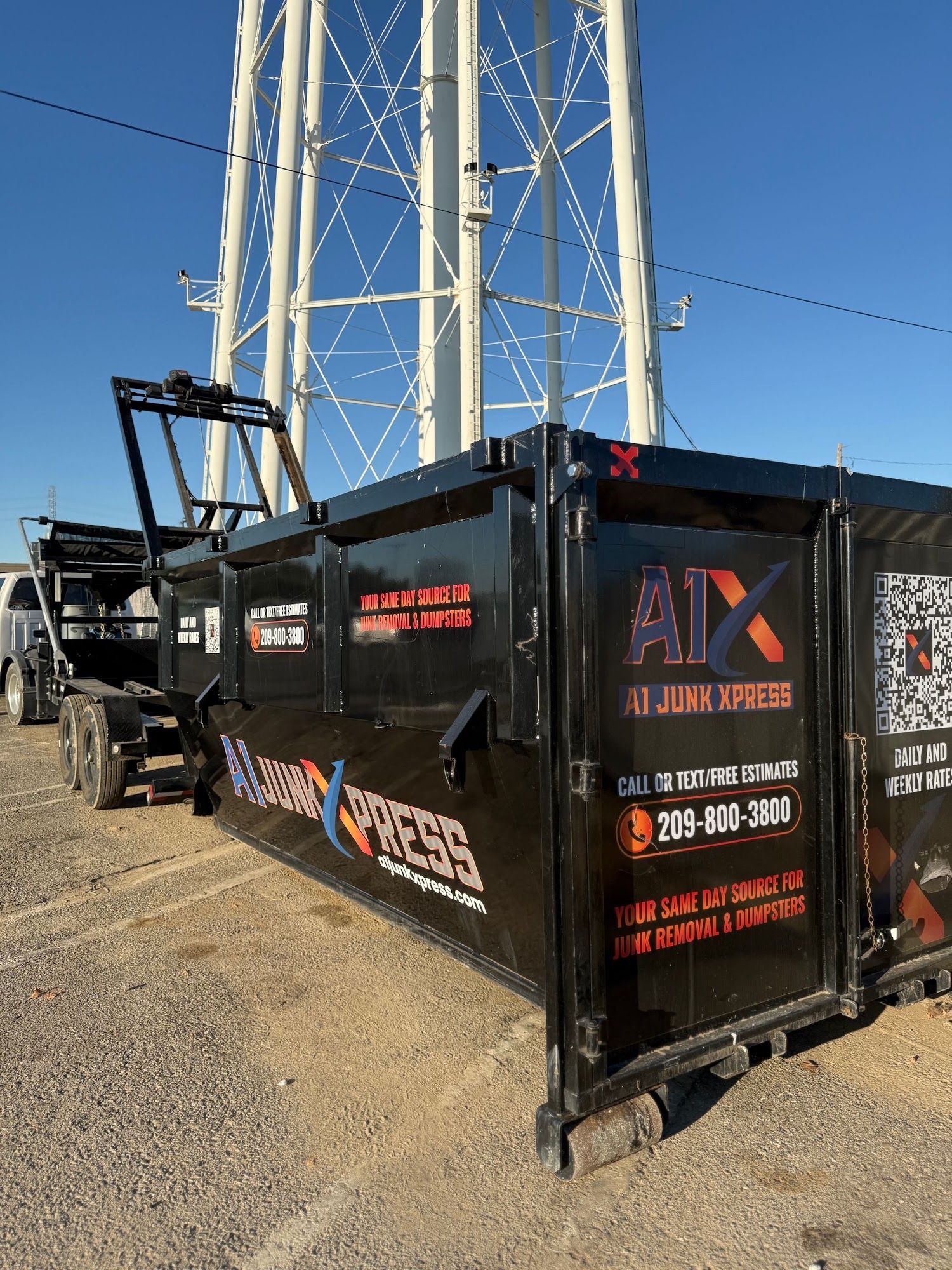 Black dumpster trailer with red logo beside a tall white metal tower on a gravel lot.