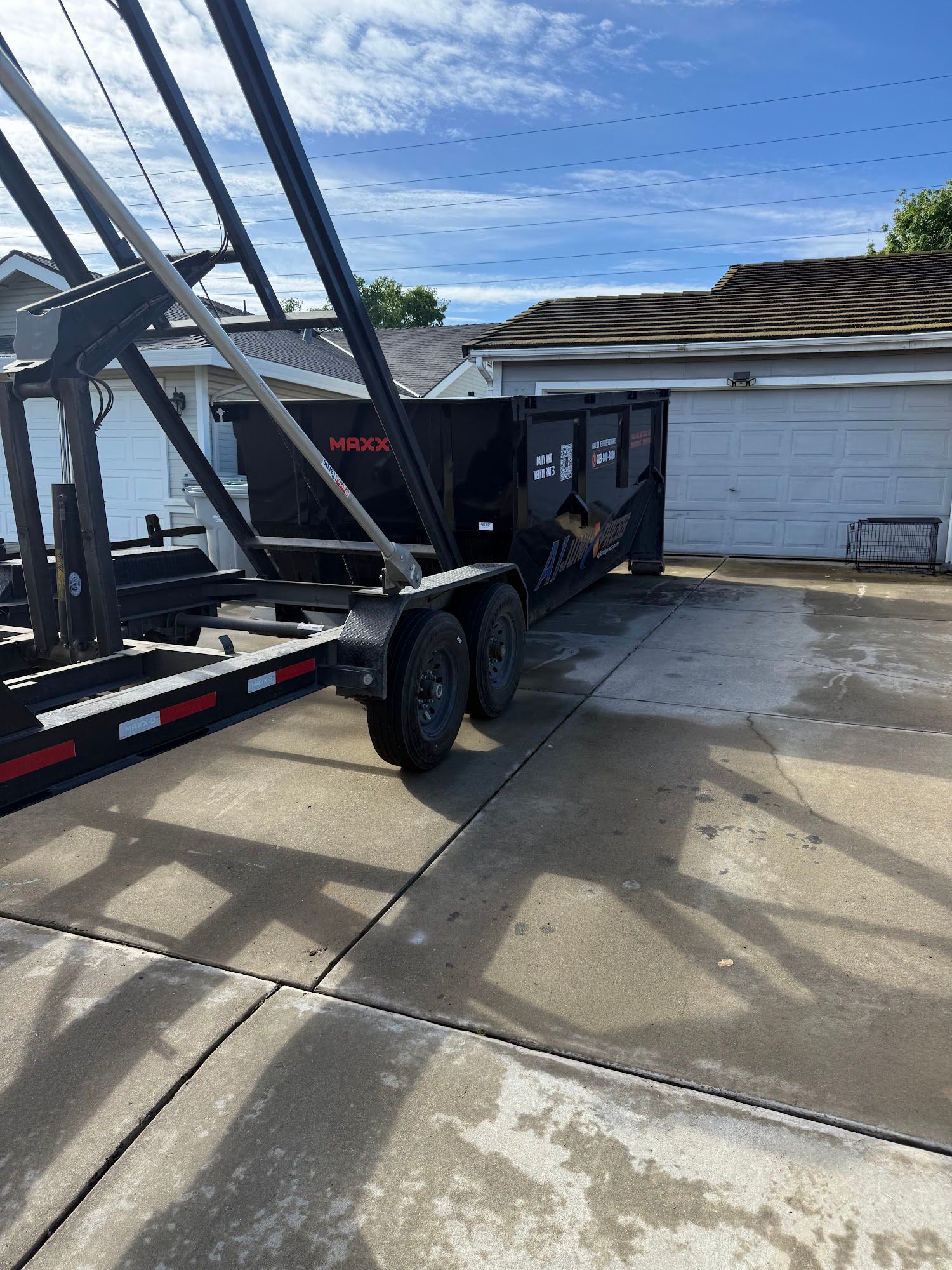 Black dump trailer parked on a wet driveway beside a white garage under a partly cloudy sky