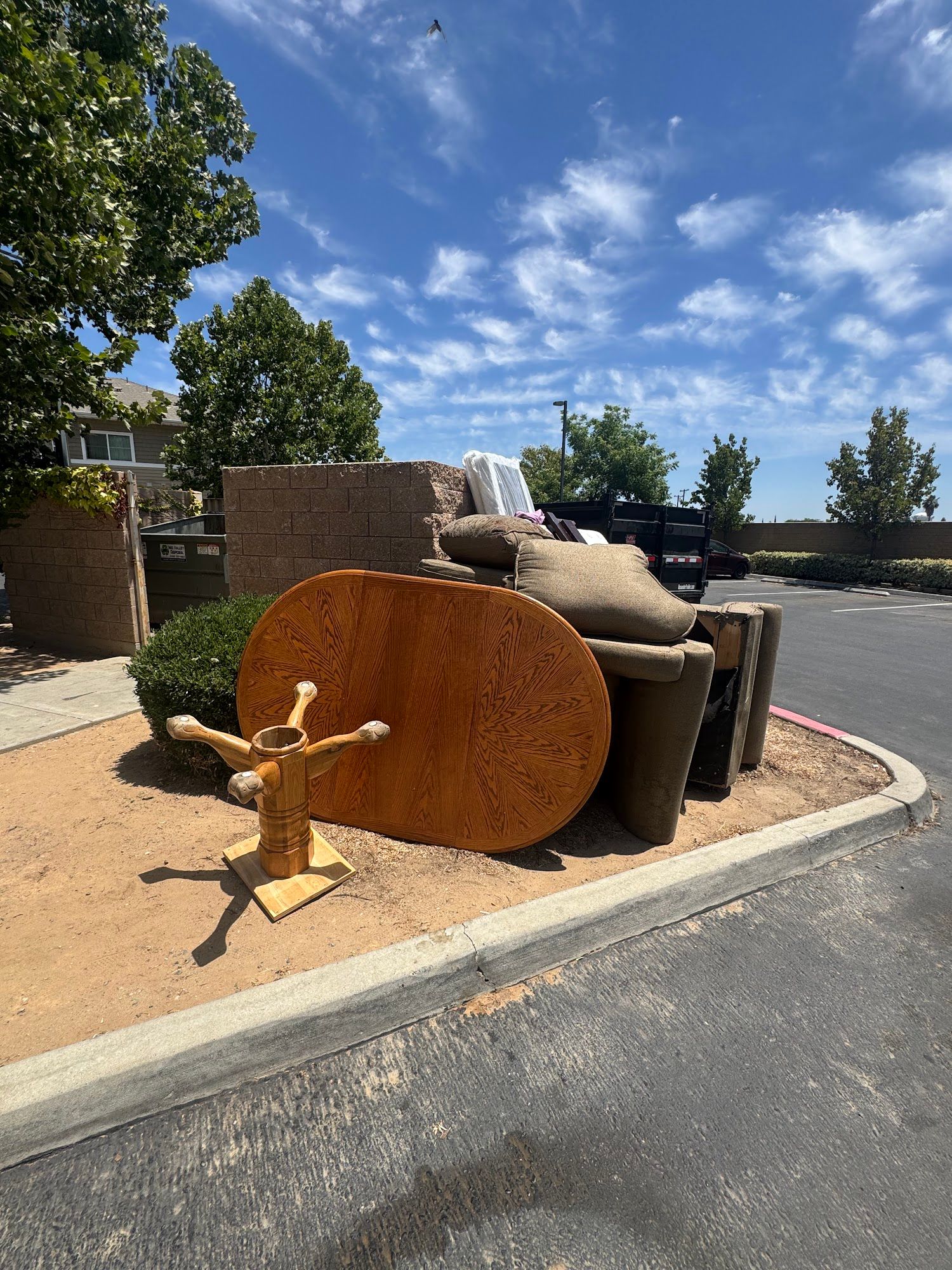 Large wooden sculpture beside a curb in a sunny parking lot with trees and blue sky