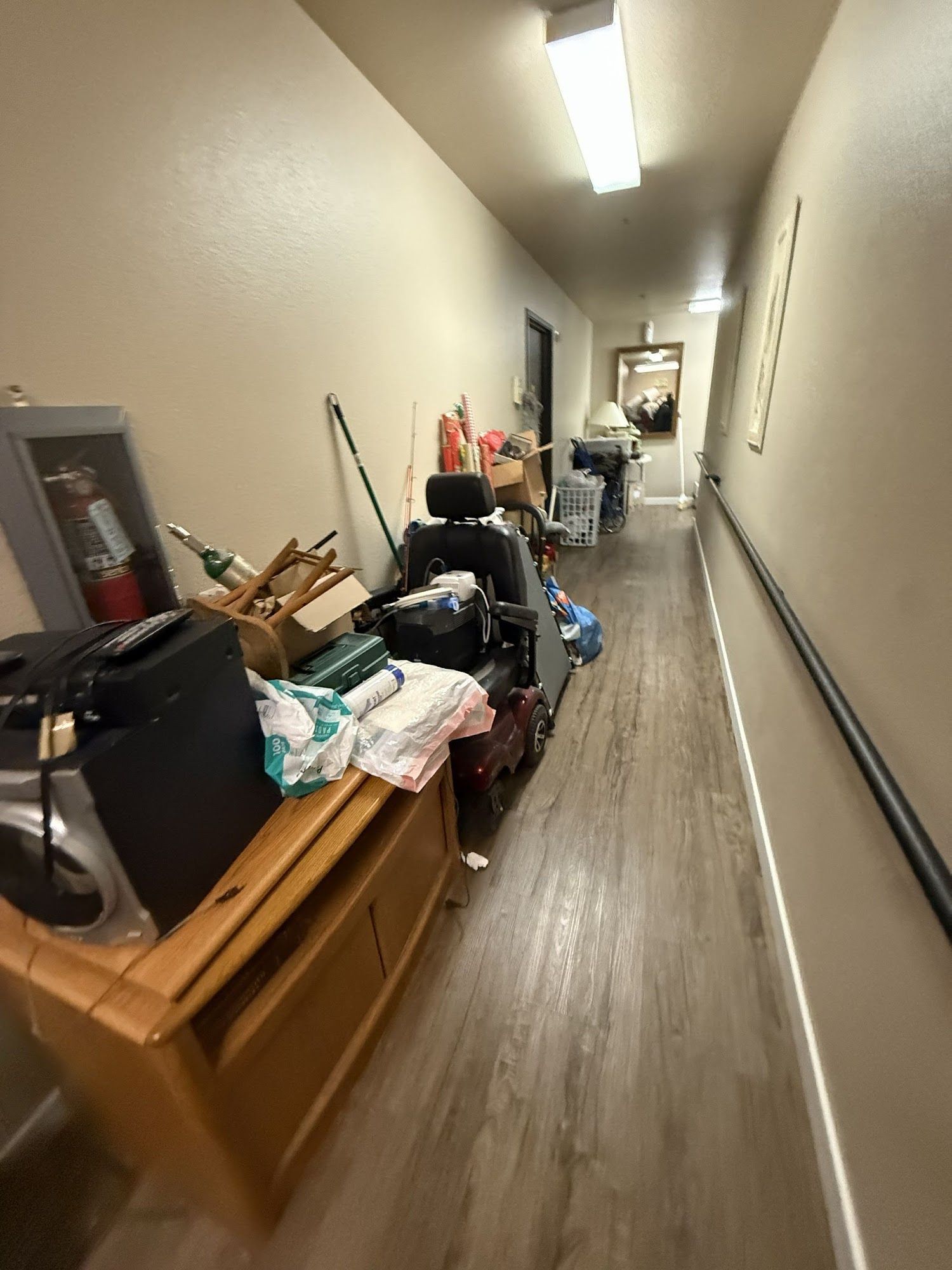 Cluttered hallway with boxes, furniture, and household items stacked along the left wall under bright ceiling lights.