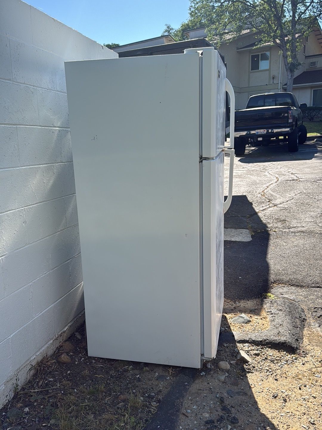 White refrigerator/freezer standing outdoors beside a cinderblock wall, with a black pickup truck in the background