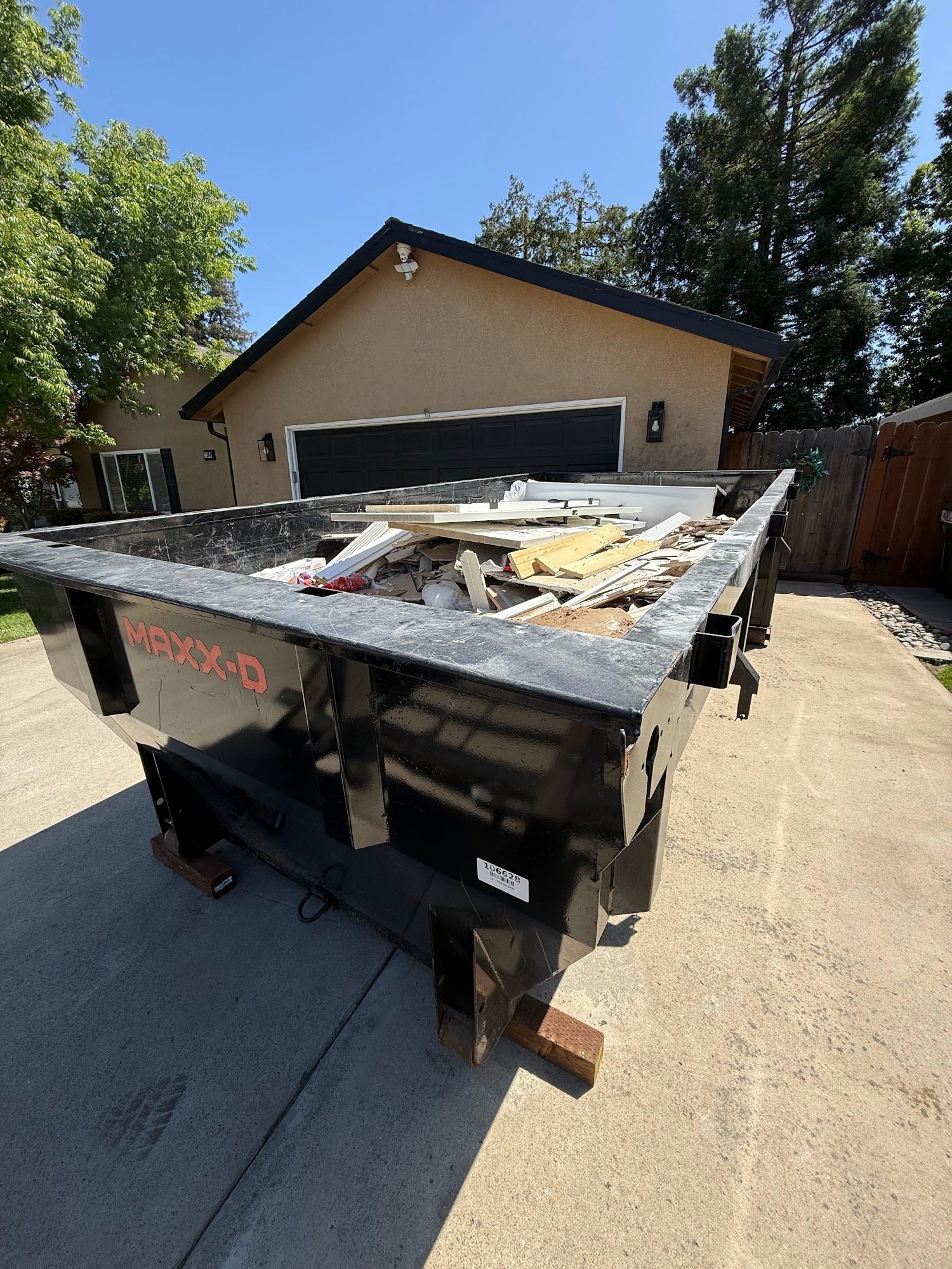 Large construction dumpster filled with debris in a driveway beside a tan house and wooden fence
