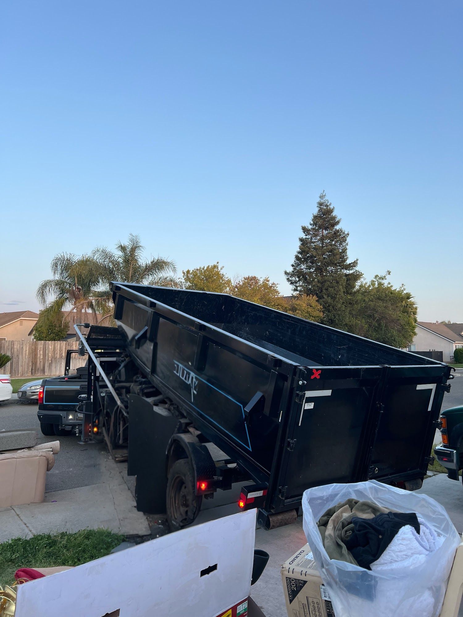 Black dump trailer tilted on a sloped driveway, with a pickup truck beside it and a clear blue sky.
