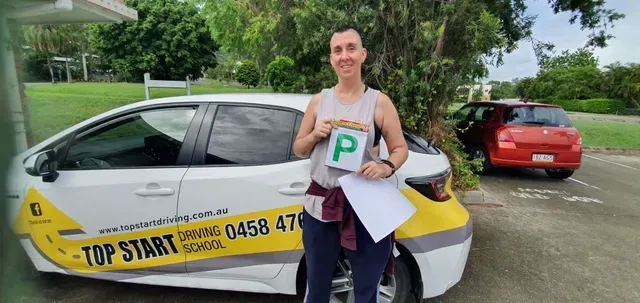 Person holding a P-plate with a car in front of a driving school car. Setting is outdoors, daytime.