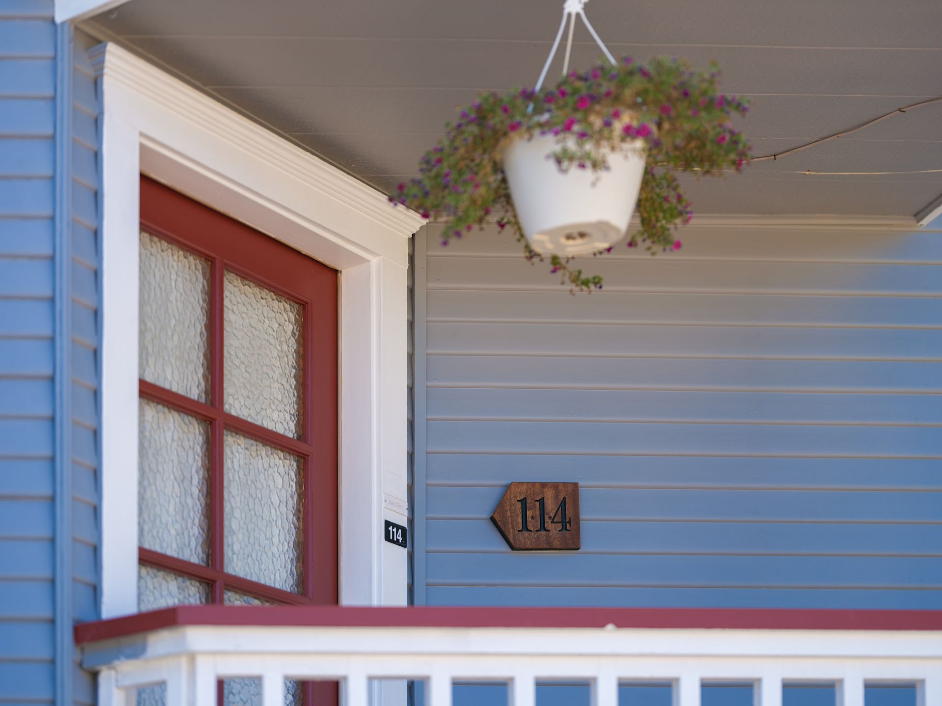 Front porch with a red door, blue siding, hanging flower basket, and house number 114.