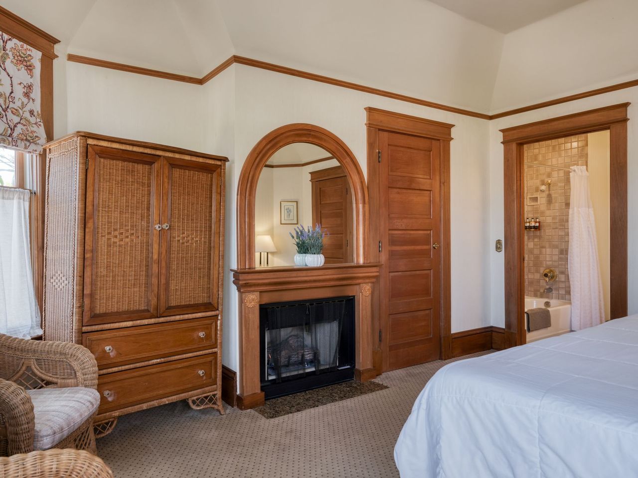 Bedroom with wood furniture, arched mirror over a fireplace, wicker cabinet, and an open doorway to a tiled bathroom