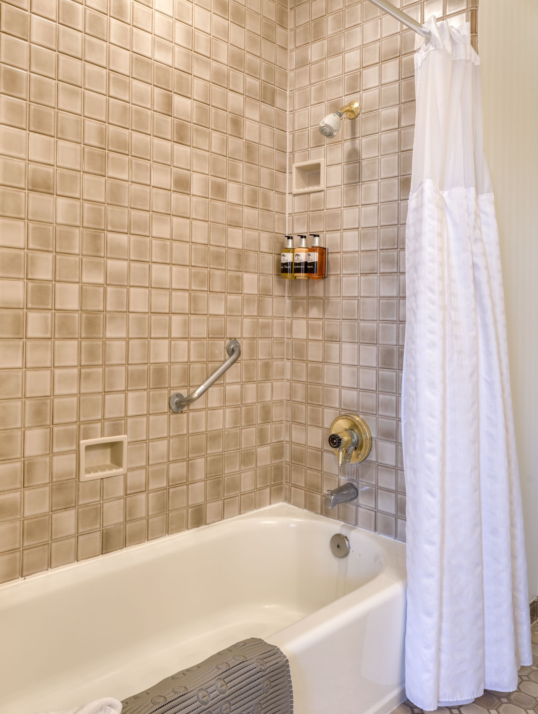 Tiled bathroom with white bathtub, grab bar, showerhead, and white shower curtain