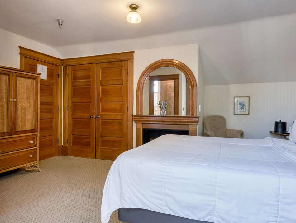 Bedroom with white bedspread, wooden closets, dresser, and arched mirror fireplace surround