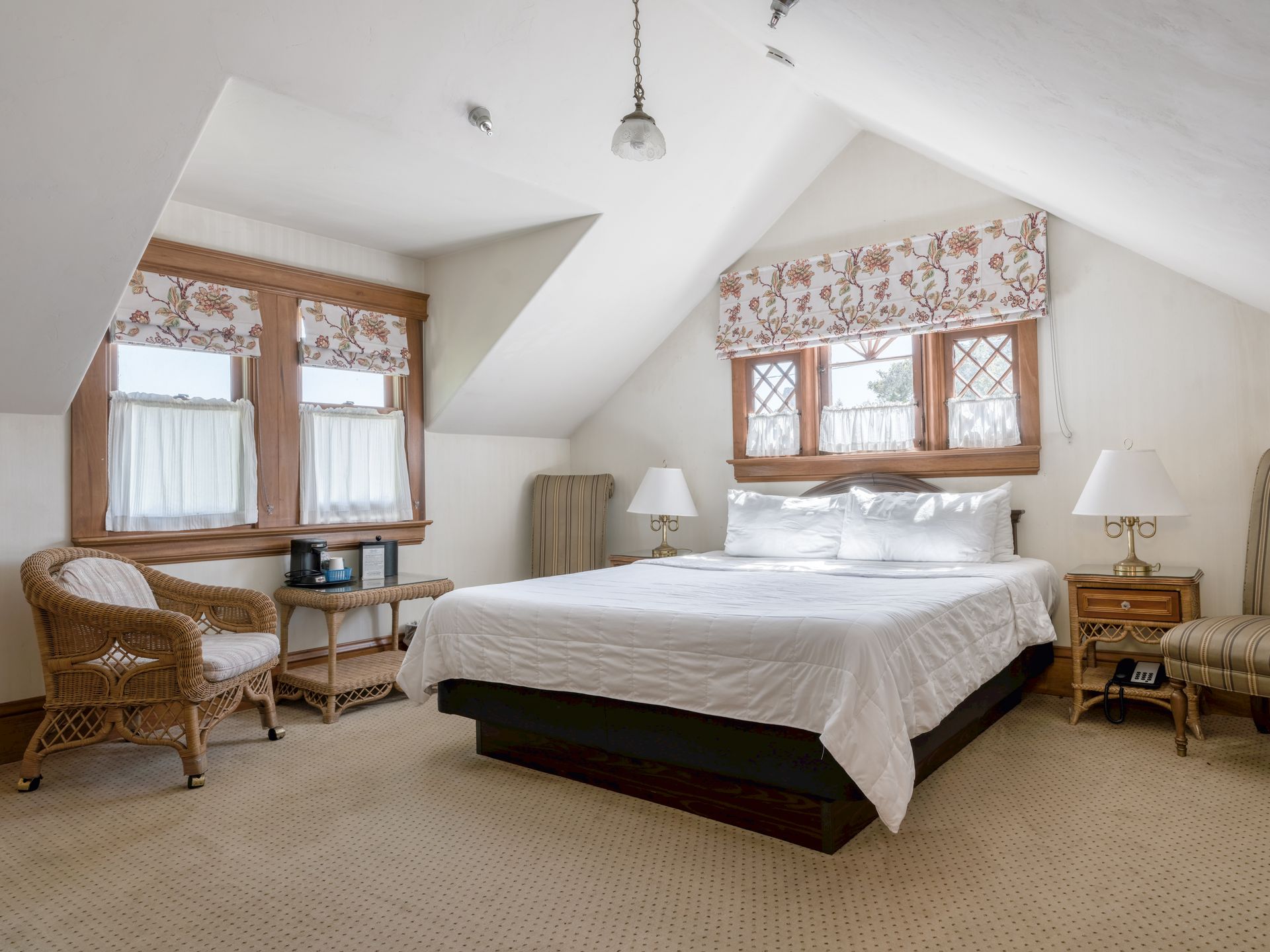 Bright attic bedroom with a bed, wicker chair, desk, and floral window valances