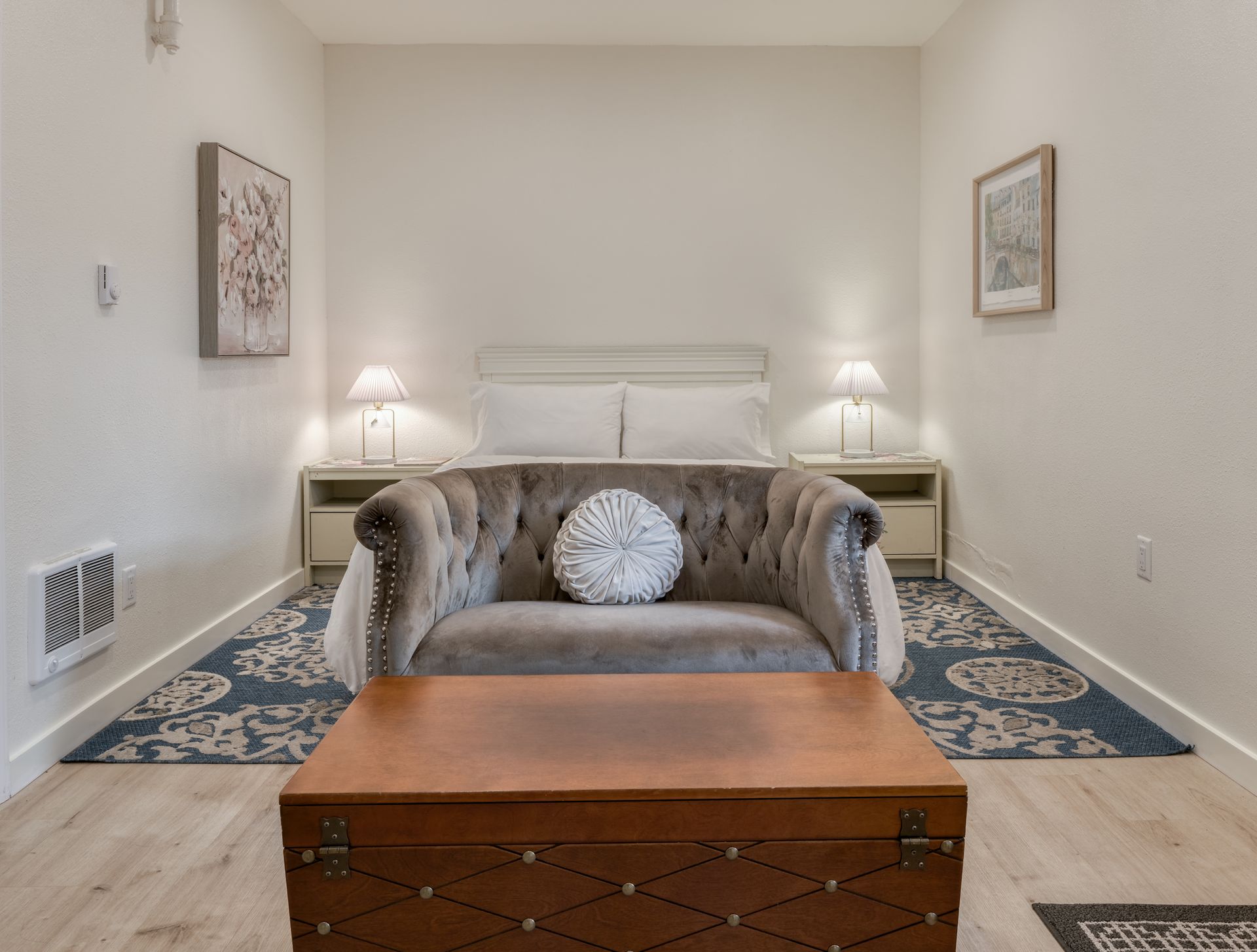 Bright living room with white bed, gray loveseat, patterned rugs, and a wooden chest in the foreground