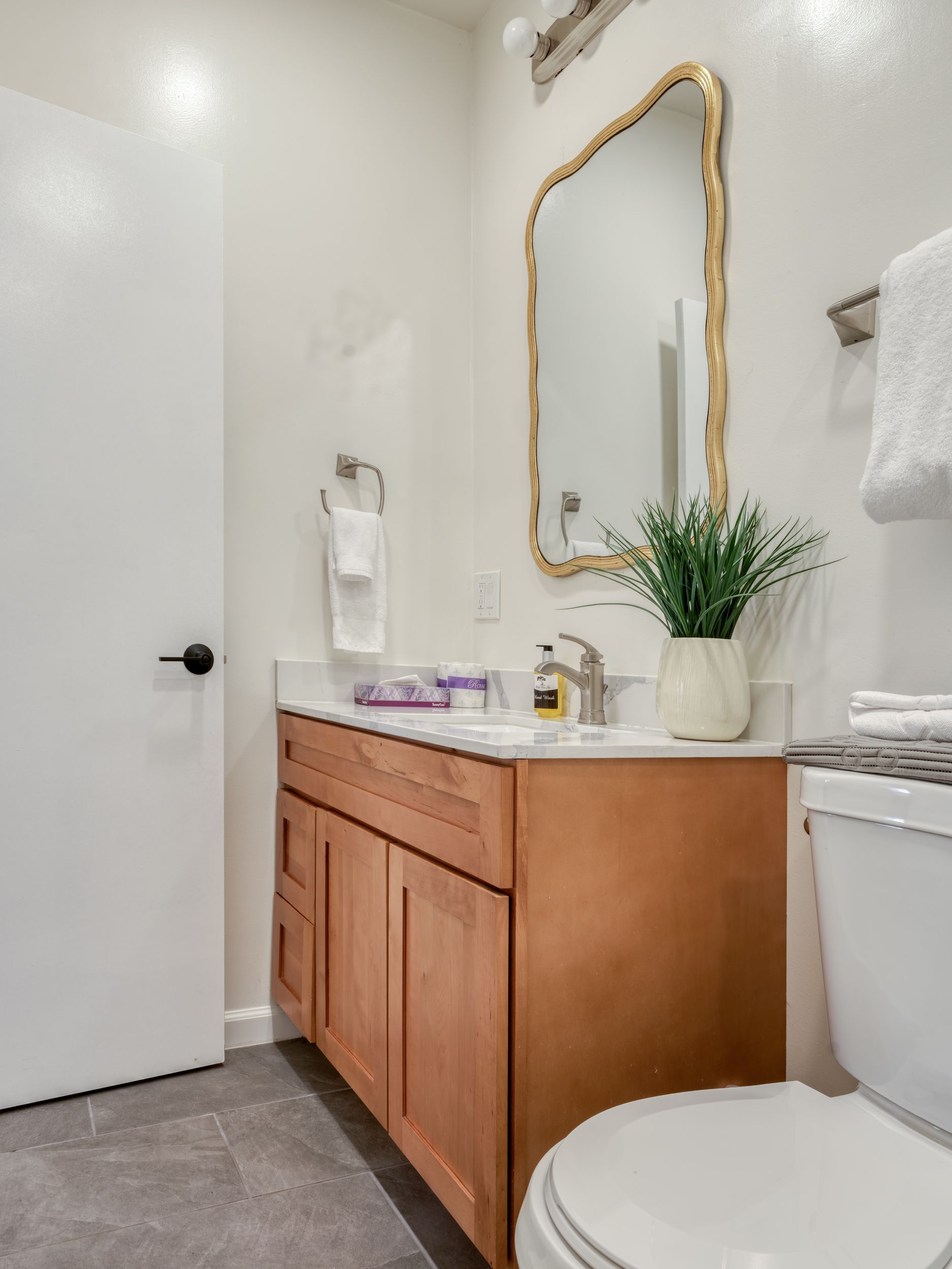 Modern bathroom vanity with gold-framed mirror, wood cabinet, white sink, and gray tile floor