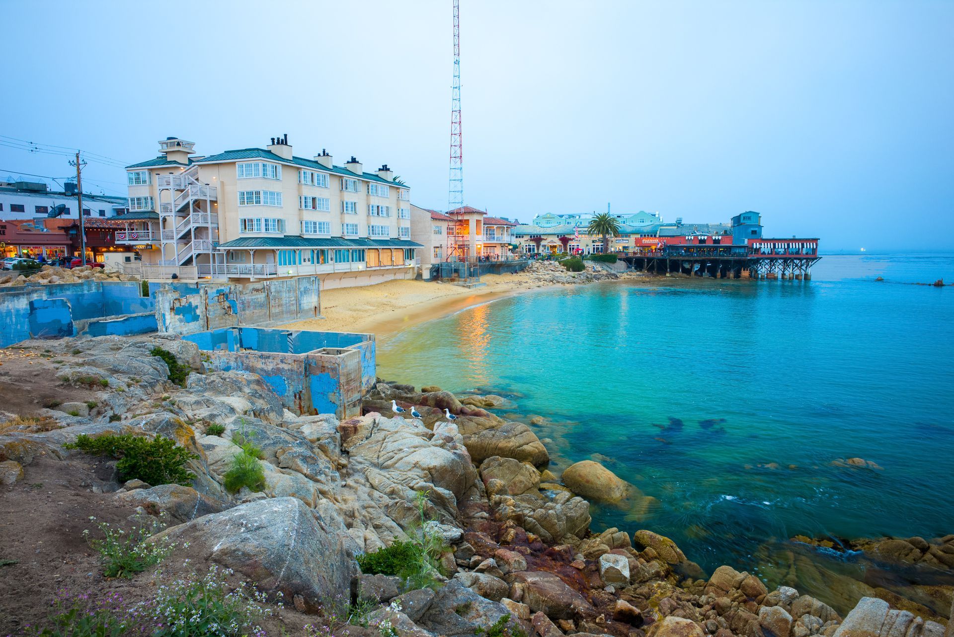 Rocky seaside shoreline with turquoise water, white buildings, and a waterfront promenade at sunset