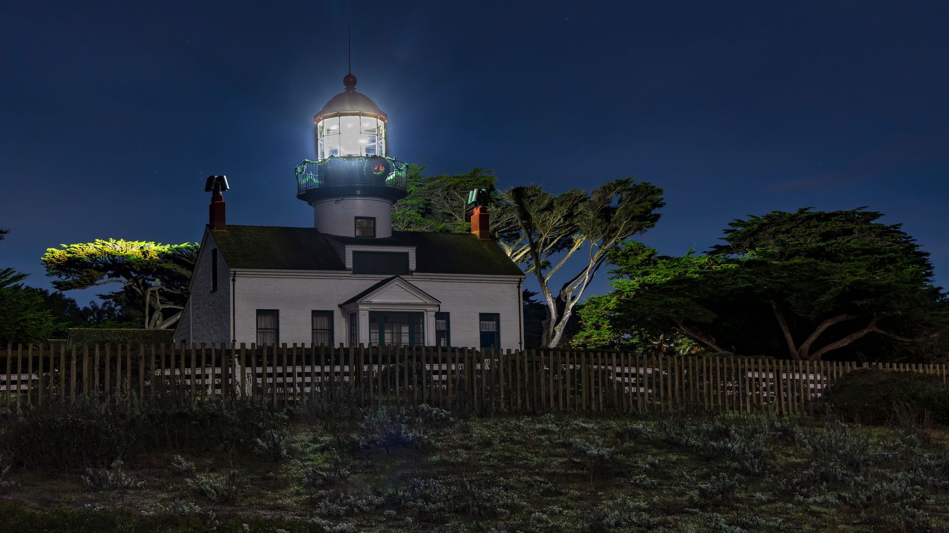 Nighttime lighthouse with white keeper’s house behind a fence, lit against a dark sky.
