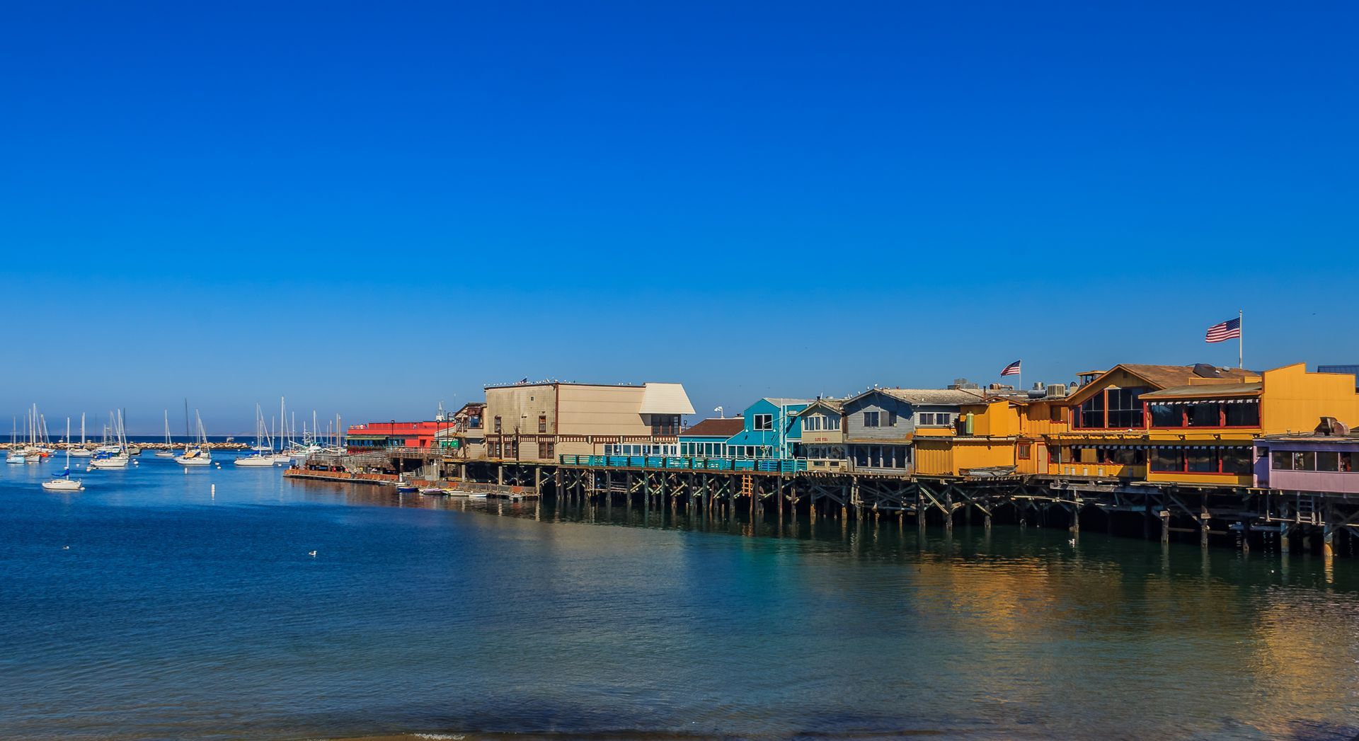 Colorful waterfront buildings on a pier overlooking calm blue water and a marina with sailboats under a clear sky