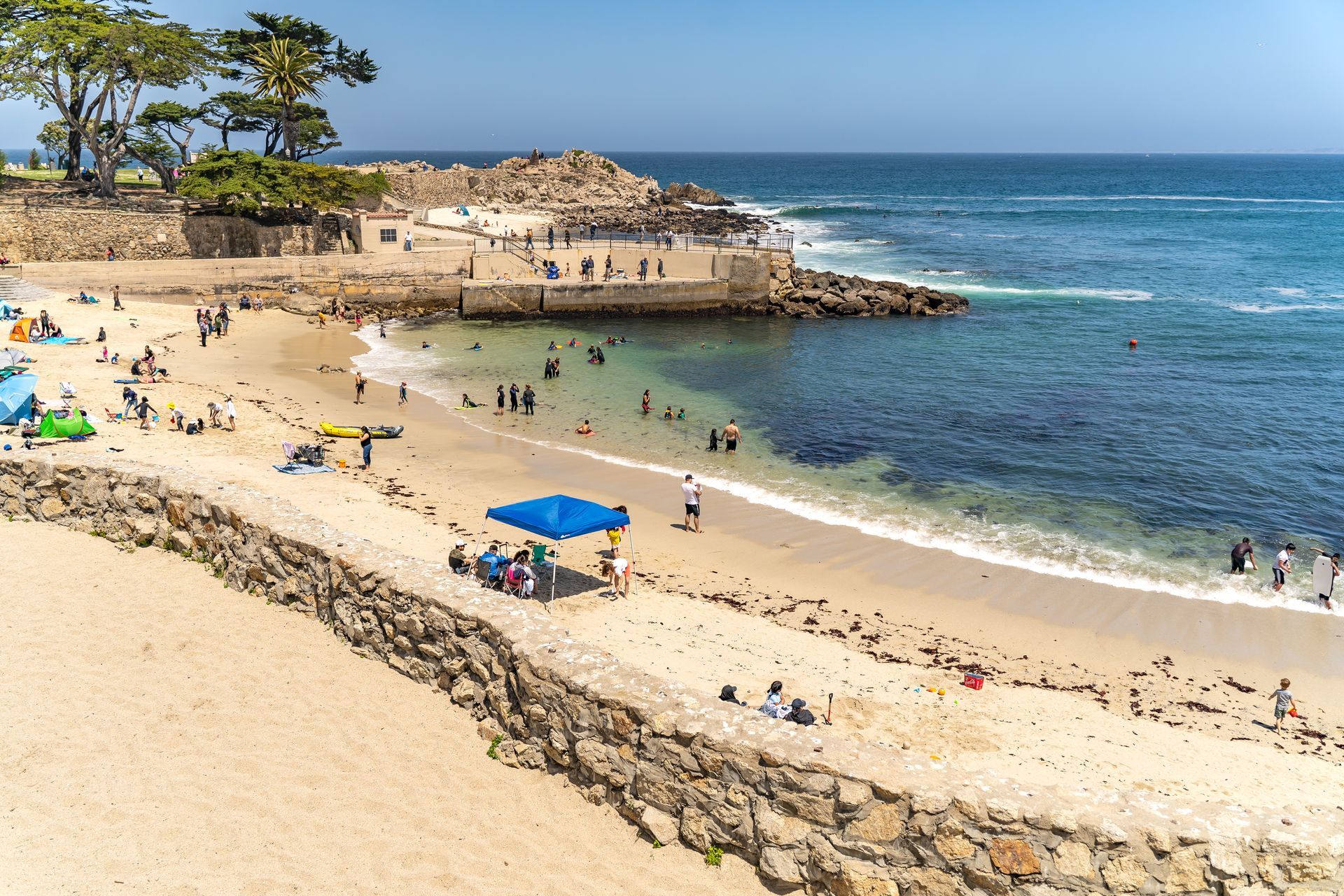 Sunny beach with swimmers, blue umbrellas, and a rocky headland beside clear ocean water