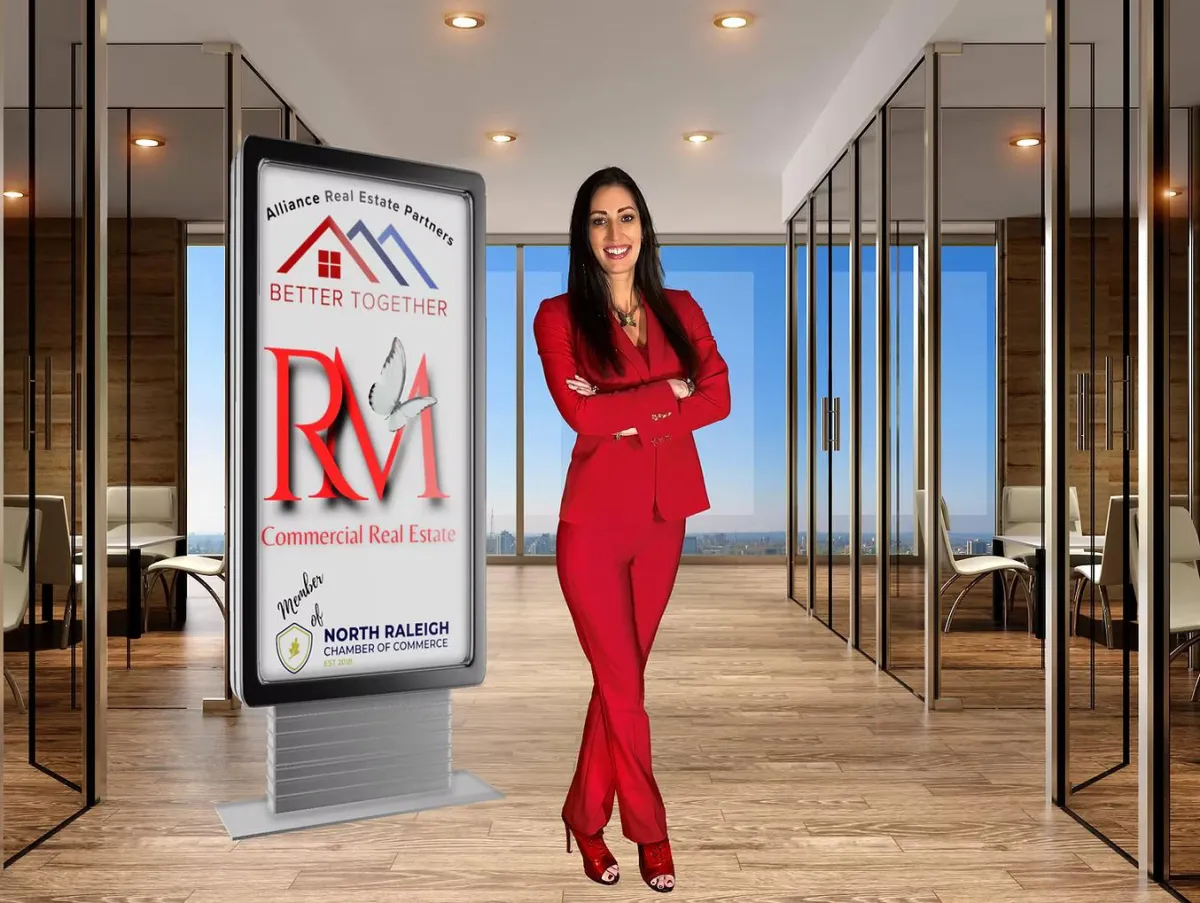 Woman in a red suit standing in a modern office lobby beside a real estate sign.