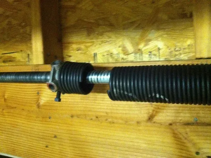 Garage door torsion spring and cable on a wooden wall, with a black corrugated cover.