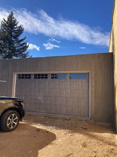 Black car beside a tan garage door under a blue sky with a pine tree and dirt driveway