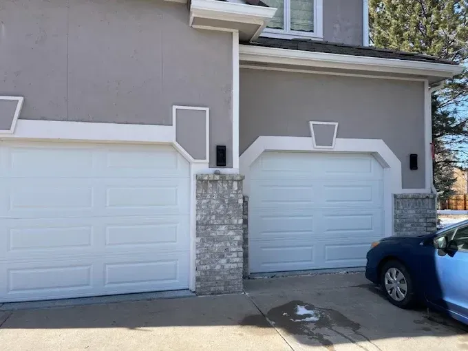 Two white garage doors on a beige house, with a blue car parked at the right edge.
