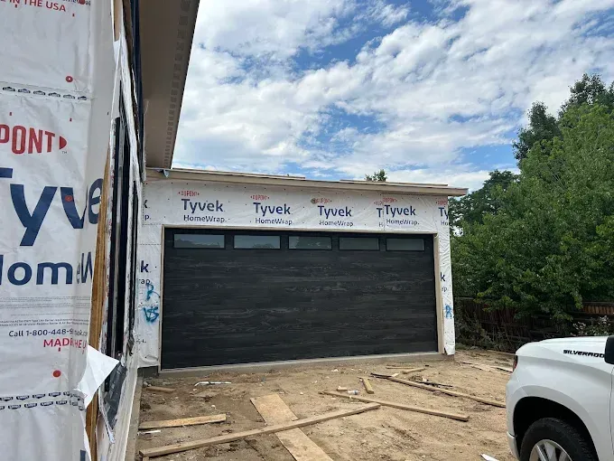 Partially finished garage with black door, Tyvek-wrapped walls, and construction materials outside